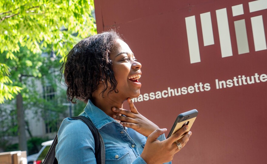 Sante, a young Black woman from Tanzania, smiles holding a phone in front of an MIT sign with white lettering on maroon background. She has short, curly, black hair and is wearing a jean shirt. She has a MIT class ring on one of her fingers.