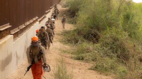 Marines with 1st Combat Engineer Battalion, armed with rifles and chainsaws, some dressed in blaze orange helmets and pants, walk in a column along the border fence move into position along the southern border to conduct vegetation clearance operations near Yuma, Ariz., Dec. 18, 2025.