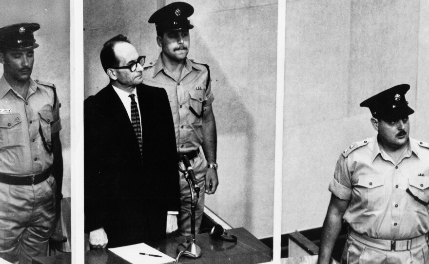 Adolf Eichmann, standing in a glass booth and flanked by guards, in the Jerusalem courtroom during his trial in 1961 for war crimes committed during World War II.