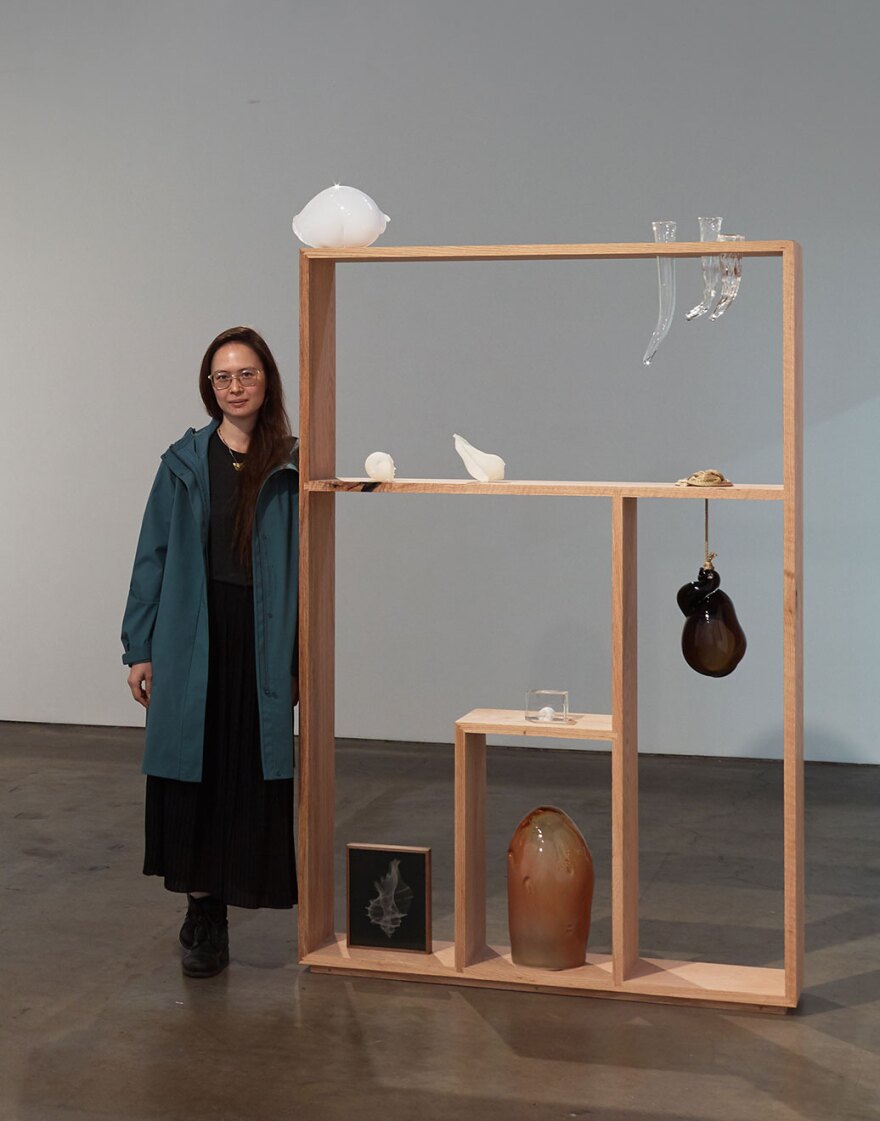 The artist stands next to a wooden display case featuring several of her glass, rope and photography works. She is wearing a long black dress and long blue-green coat, and glasses. She has long reddish-brown hair.