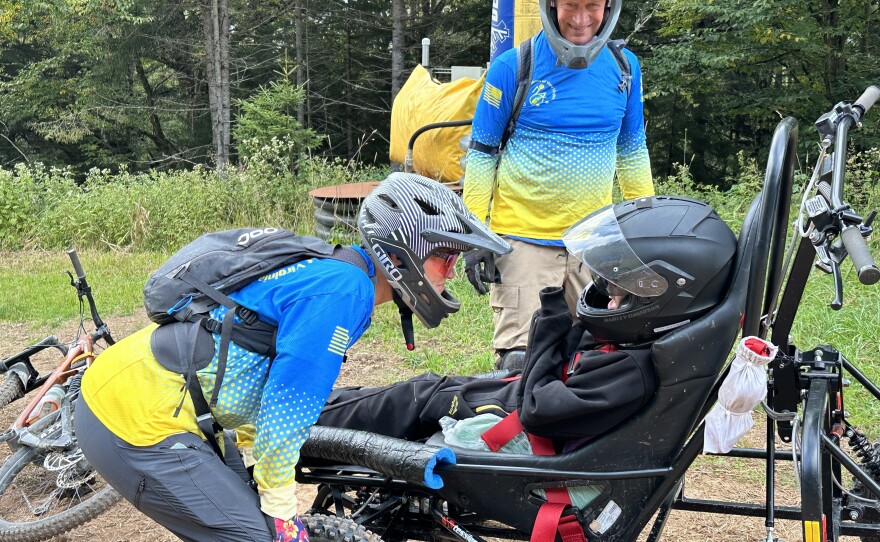 Carol Woody leans forward with her hands on her knees as she asks Gage Tatar if he's ready to ride again in the Cimgo. Gage Tatar smiles from underneath his mountain biking helmet while a volunteer, Erwin Berry stands by ready to help.
