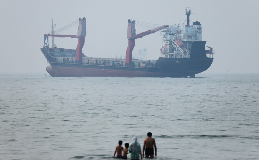 A group of people stands in shallow water as a cargo ship appears anchored in the Strait of Hormuz off Bandar Abbas, Iran, Tuesday, June 30, 2026.