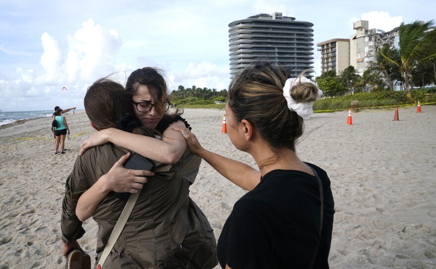 Faydah Bushnaq, of Sterling, Va., center, is hugged by Maria Fernanda Martinez, of Boca Raton, Fla., as they stand outside the partially collapsed building on Friday. Bushnaq is vacationing and stopped to write "Pray for their Souls" in the sand.