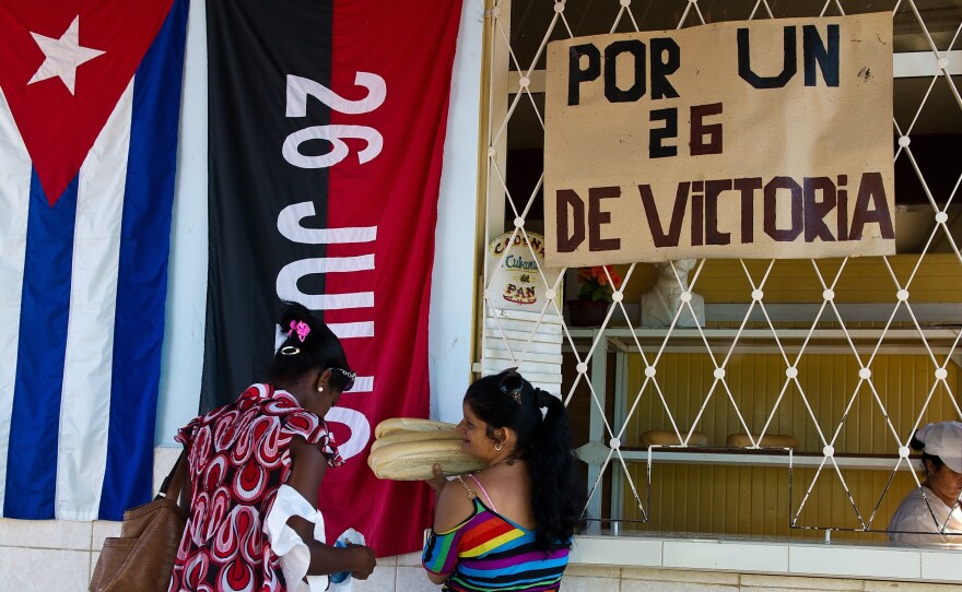 The flag of the July 26 Movement hangs to the right of the Cuban flag, behind two women buying bread in the city of Artemisa.