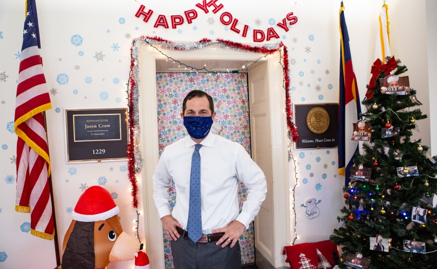 Rep. Crow stands outside his office door, with the tree filled with photos of Rep. Perlmutter behind him.