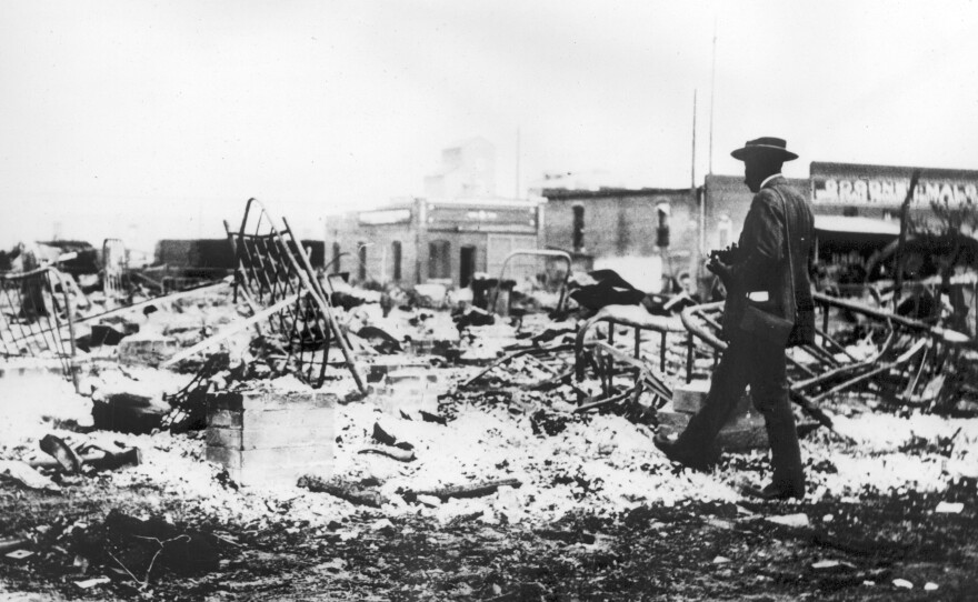 Photograph of an African-American man with a camera looking at the skeletons of iron beds which rise above the ashes of a burned-out block after the 1921 Tulsa Race Massacre, in Oklahoma.