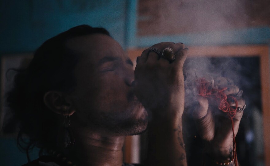 A close up of brown-skinned person with head slightly tilted back and eyes clasped shut while smoke billows around them. Hands are raised in front of their face as if smoking; the left hand clutches an object with orange yarn danging down, the right hand features two rings and partially obscures the object. The background features an interior space with blue walls.