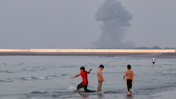Three boys play in the shallow waters of the Strait of Hormuz, as a plume of smoke rises from an explosion in the background, off Bandar Abbas, Iran, Monday.