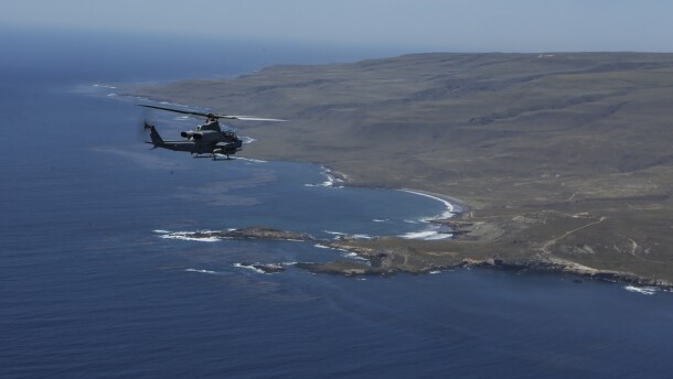 A Marine Corps Ah-1Z SuperCobra attack helicopter flies from the left toward the western rocky shore of San Clemente Island.