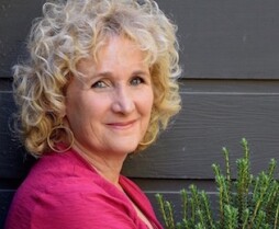 Headshot of Nan Sterman wearing a pink shirt, standing in front of gray wall, next to green plant