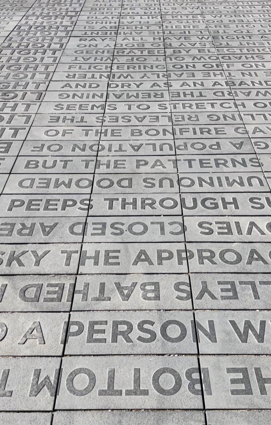 Hundreds of seemingly disconnected words are carved on concrete in an outdoor walkway art installation. The words appear dark gray against a lighter gray backdrop. Visible are words like "BOTTOM," "PERSON" "DOOMED" "PEEPS THROUGH"' and many more. The words are in all caps and vary in direction and whether they're upside down. The concrete is divided into rectangular pavers. The walkway fills the full photo.