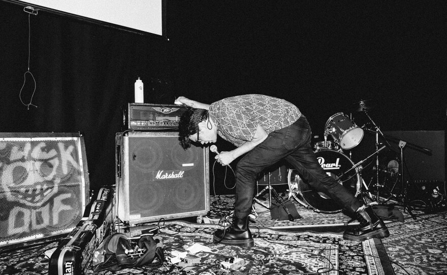 A male singer is shown in a black and white photo lunging forward, leaning over the mic, and steadying himself against a large Marshall amp. He's on a stage covered in decorative Persian-style rugs, with an empty drum set behind him.