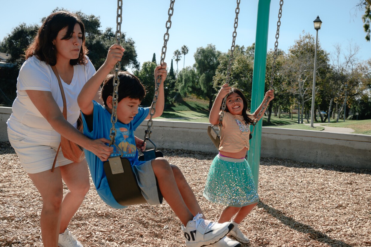 Alondra Padilla pushes her son, Mateo, while her daughter, Valentina, watches on the swings at Hilltop Park in West Chula Vista on Oct. 31, 2023.