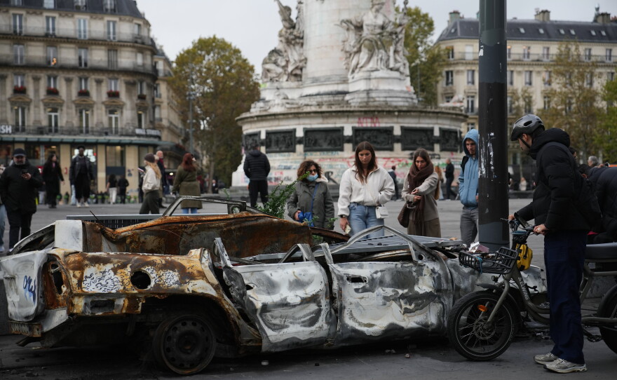 People watch a burned-out car after a quiet night Monday under curfew, the first since weekend protests over the cost of living turned into rioting and looting across France, in Paris, Tuesday, Oct. 20, 2026.