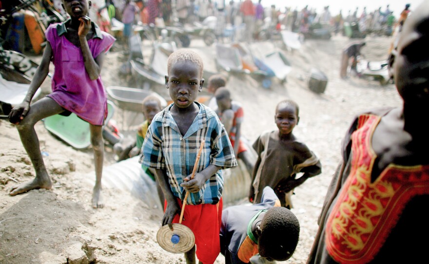 A boy holds a makeshift toy. Behind him, young men wait with wheelbarrows for food rations to be distributed.