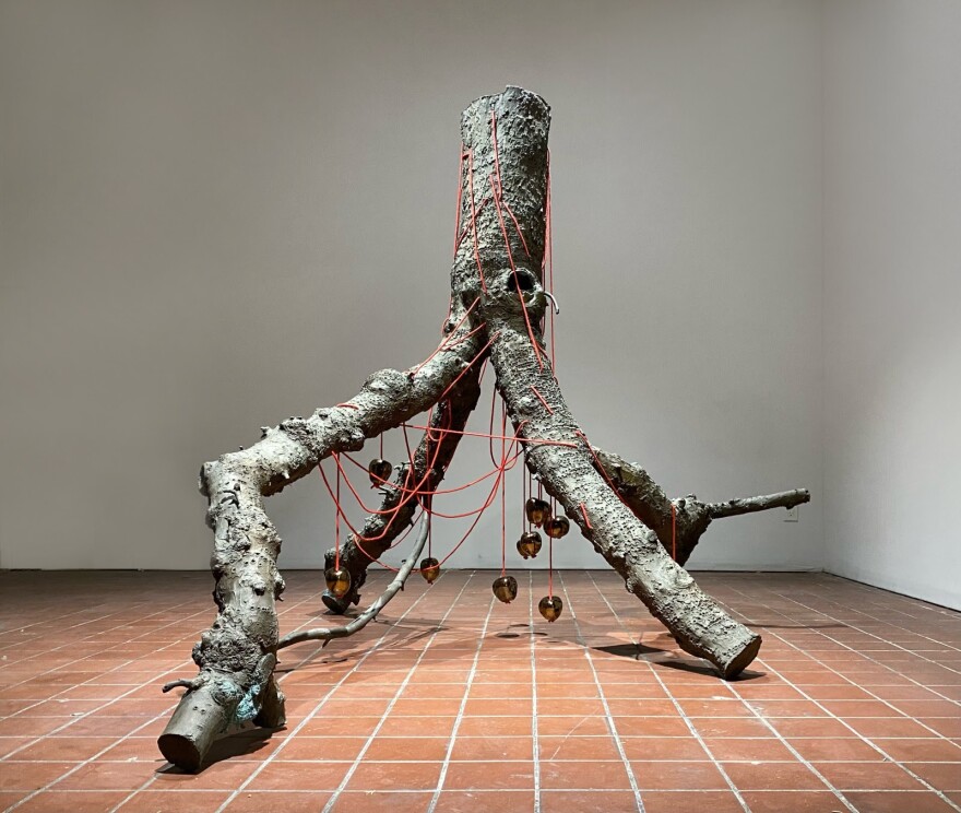 Anya Gallaccio's "Rêvons D'or" sculpture is a large, life-sized bronze cast tree trunk and branches, positioned upside down, with porcelain apples hung by a red rope wrapped around the trunk and branches.