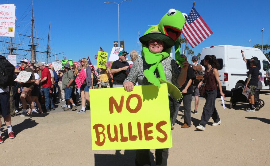 Mary Decker is wrapped with a Kermit the Frog plush toy and holds a sign that reads "No Bullies" during the "No Kings" protest rally at Waterfront Park on Oct. 18, 2025.