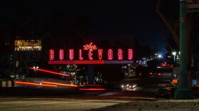 A nighttime street scene featuring a large neon sign that reads "the BOULEVARD" in bright red letters. The sign is illuminated against a dark sky, with blurred car lights streaking past on the road below.