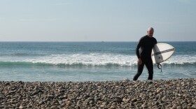 A surfer walks on the rocks between the water and the road named "The Strand" in front of Tyson Street park in Oceanside, California. Coastal erosion has washed away most of the beach's sand. The rocks serve as a replacement until the proposed project REBEACH is underway in late 2026.