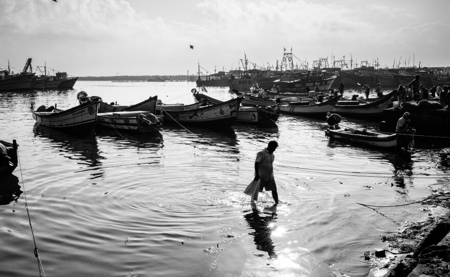 A fisherman wades to the shore from his anchored boat, as the sun rises higher in the sky and the activity on the docks winds down for the day.