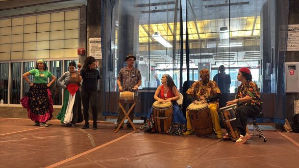 Four people sit with drums while third plays a maraca and two dancers prepare to perform.