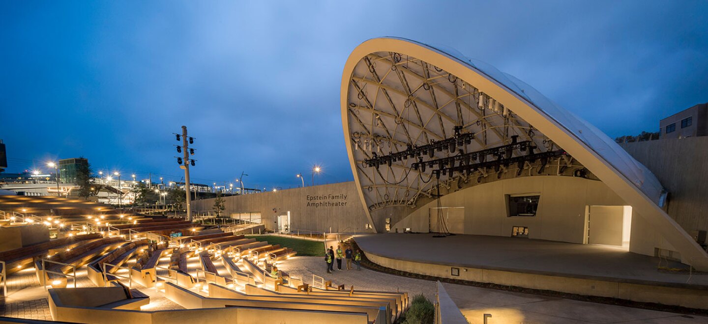 The new UC San Diego outdoor amphitheater is lit up at dusk against a dark blue sky. A curved shell-like cover provides a partial roof for the concrete stage. Concrete and wooden bench seating forms a segmented semi-circle around the stage.