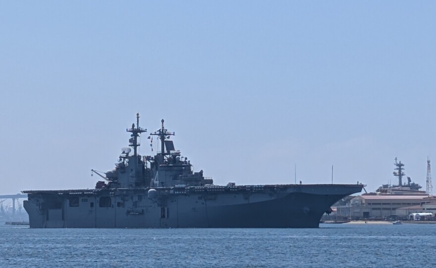 The amphibious assault ship USS Boxer transits San Diego Bay moving left to right in the photo with Naval Air Station North Island and the San Diego-Coronado Bridge visible in the background.