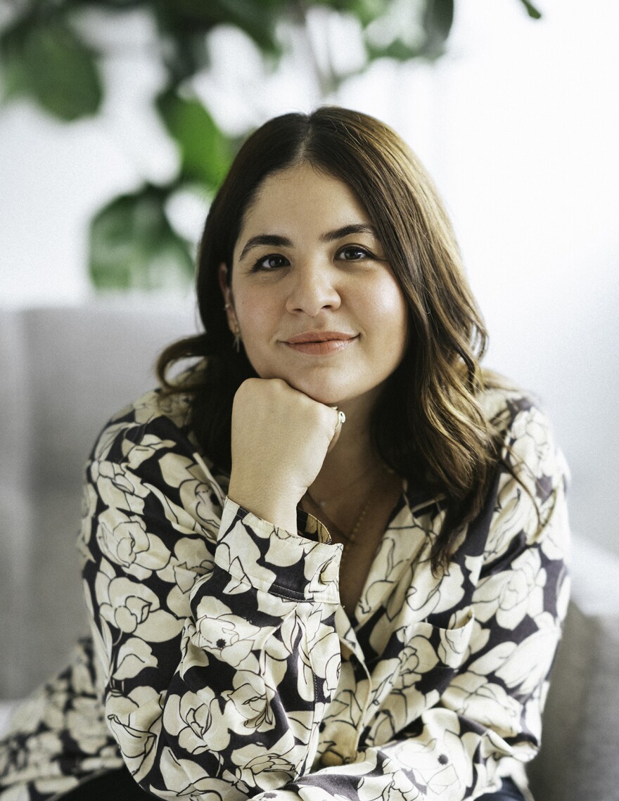 The third Poet Laureate of San Diego, Paola Capó-García, is shown in an undated photo. She is looking at the camera and smiling, resting her chin on her fist. She is wearing a black and white floral blouse, and has medium-length brown hair.