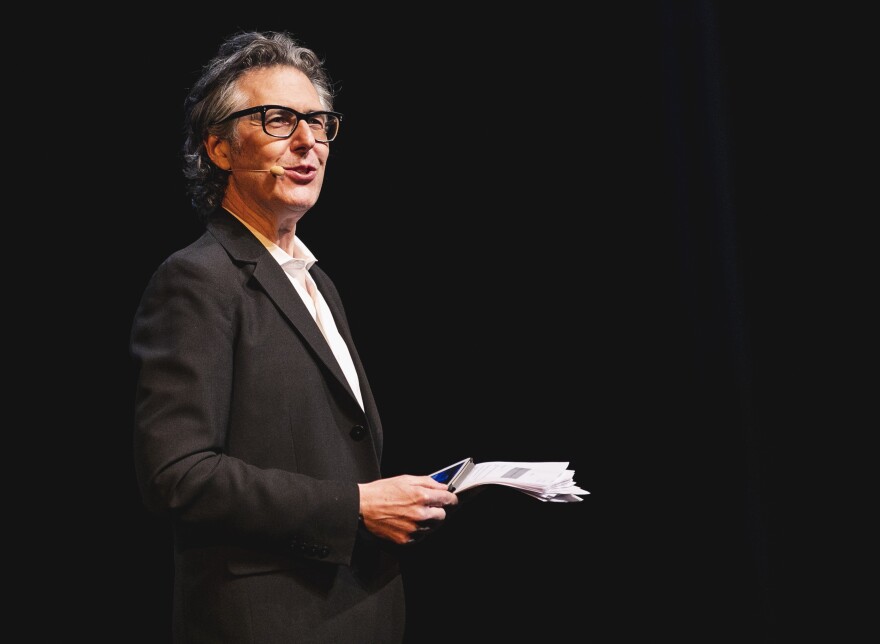 Ira Glass is shown during a live show in 2019. He is holding a stack of papers and a tablet, and wears a small headset mic.