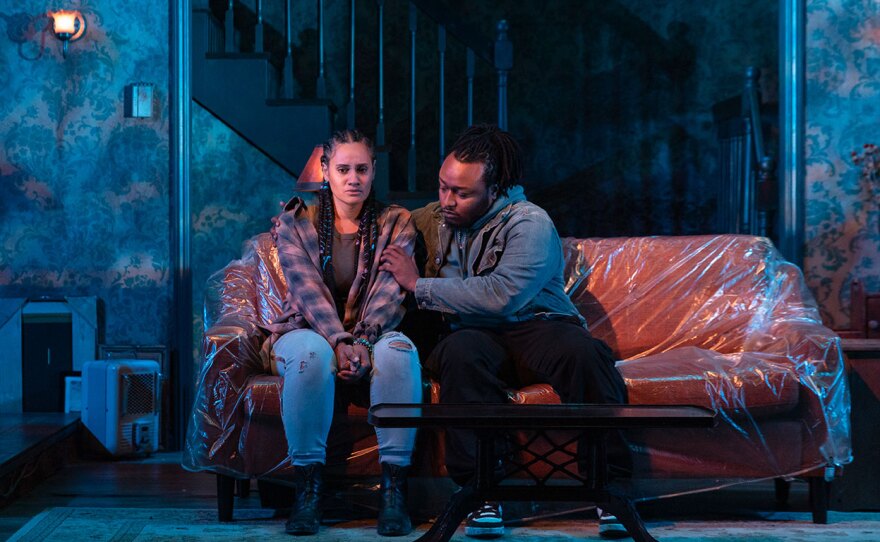 Two actors sit on a couch wrapped in plastic on Cygnet Theatre's stage.