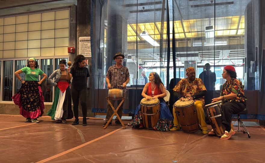 Four people sit with drums while third plays a maraca and two dancers prepare to perform.