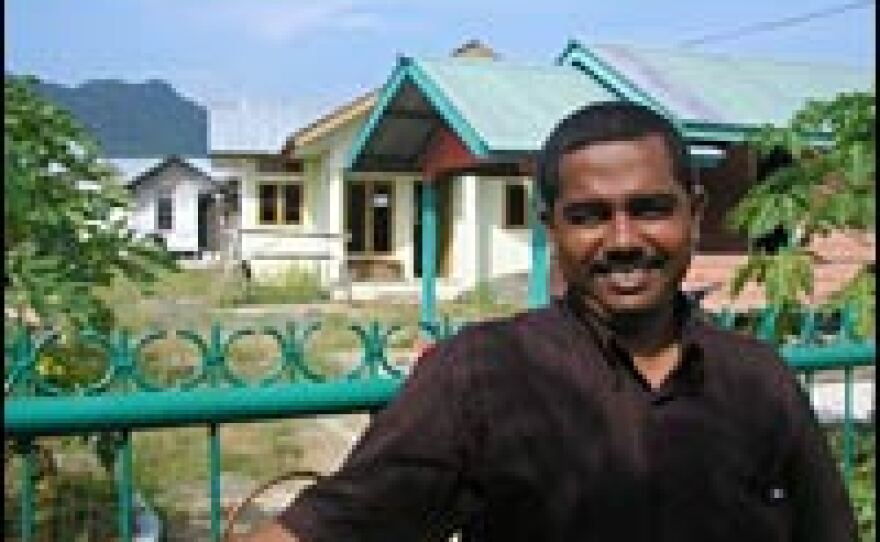 Mursalin stands in front of the temporary house he built last year on his own. Behind it is the permanent house built on his lot for him by Catholic Relief Services. His new house is almost finished, but CRS says problems with contractors' work will require them to retrofit it, a process that will take several more months.