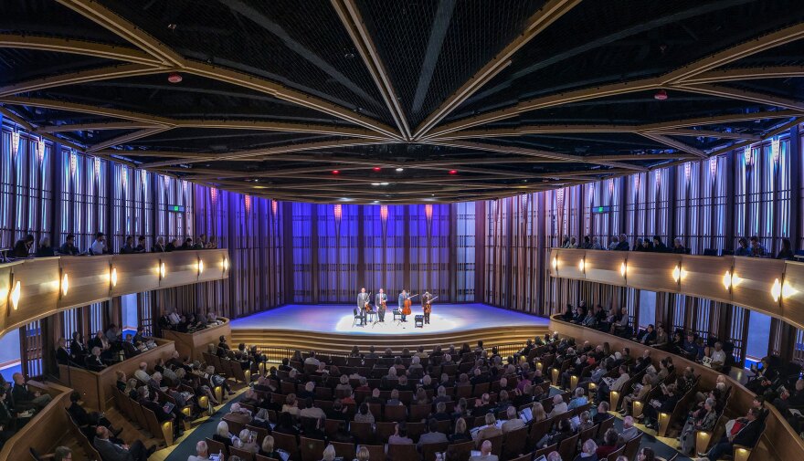 A quartet stands and bows on stage to an audience in a concert hall. The walls are illuminated in gold, blue and purple light and the ceiling has light, golden wood architectural accents.