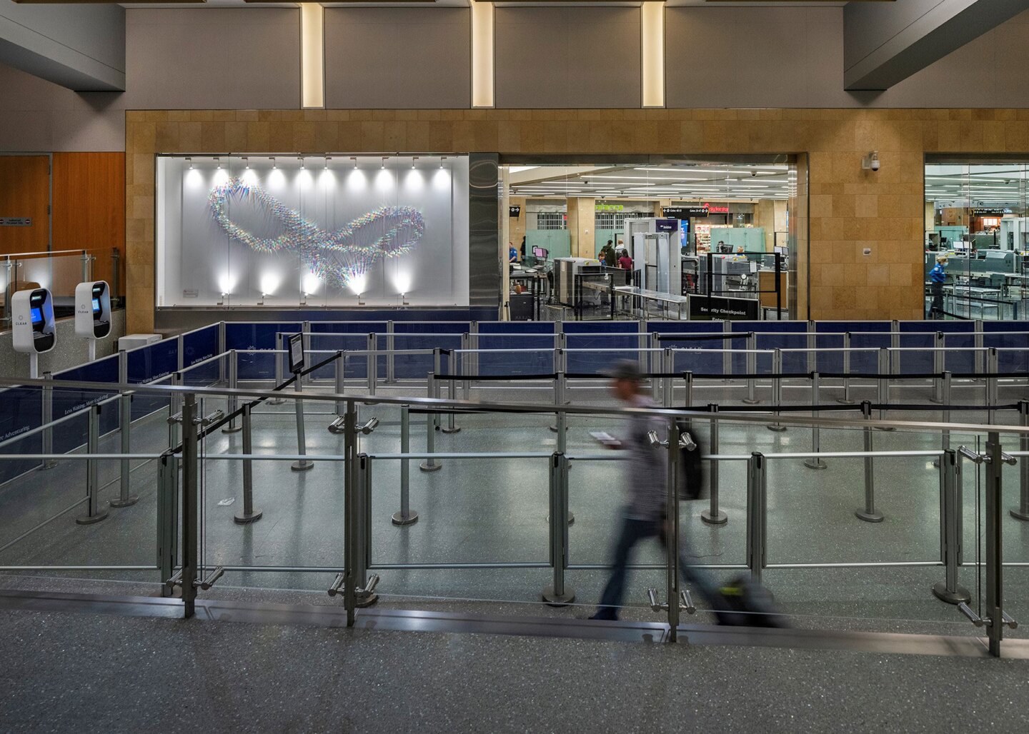 Artist Kaori Fukuyama's installation is shown in San Diego International Airport in the pre-security zone by Checkpoint 6 in Terminal 2. A traveler, shown blurry as if in motion, wheels a suitcase through the line adjacent to the glass display case. The security line is otherwise empty.