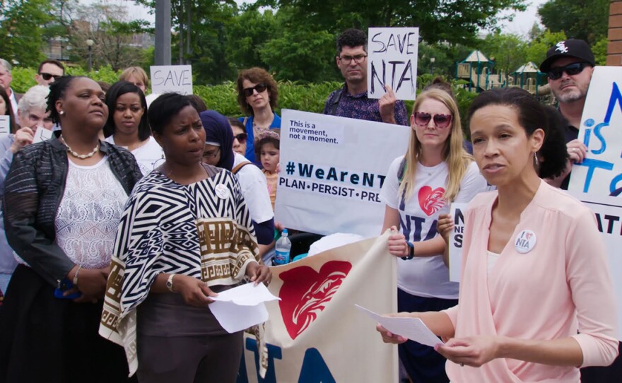 Person reads a speech at a rally. In the surrounding crowd people hold up signs saying "Save NTA" and "#WeAreNTA."