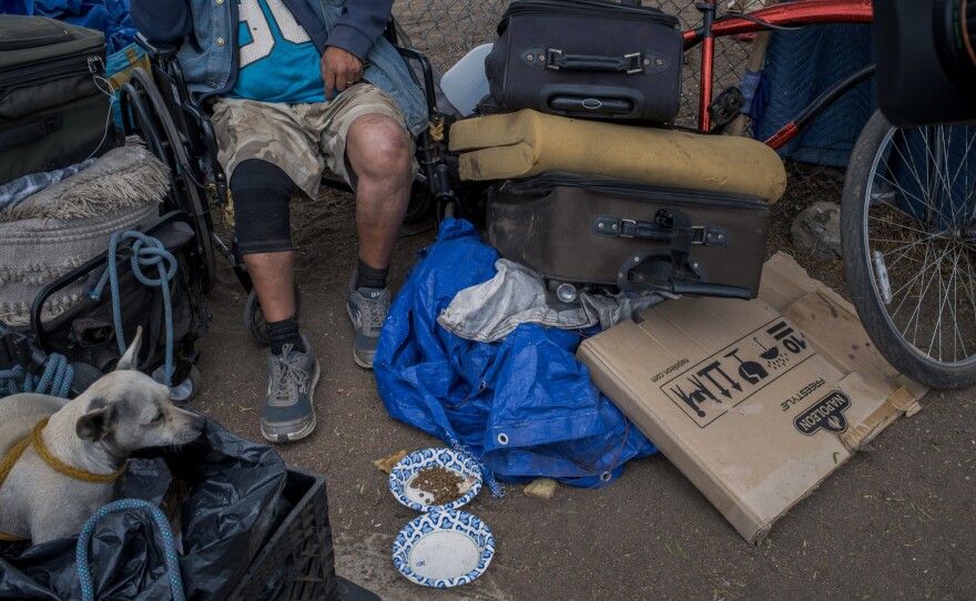 Juan Carlos Rivera and his dog sit with his belongings on the city's side of a chain link fence in San Diego on Friday, Aug. 15, 2025.