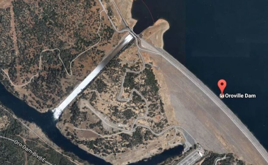 A Google Map image shows the tall Oroville Dam (lower right) with the main spillway allowing a flow of water. The auxiliary spillway is seen (at the upper left) adjacent to the main spillway.