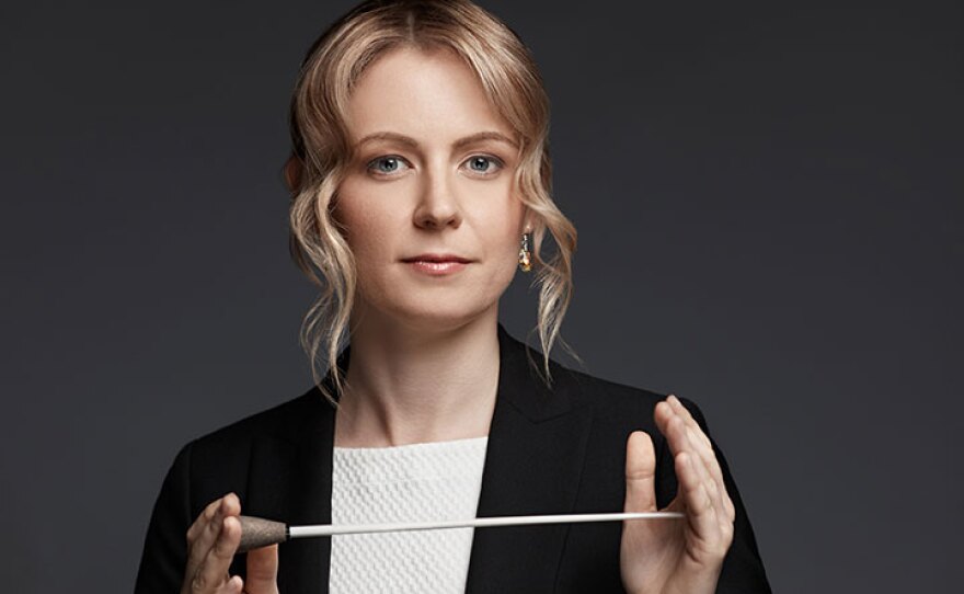 Conductor Gemma New is shown wearing a black jacket and white shirt. Her hair is blond, and curled around her face. She's holding a conductor's baton with both hands, horizontally in front of her. She is looking at the camera.