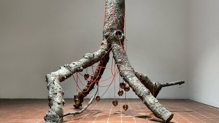 Anya Gallaccio's "Rêvons D'or" sculpture is a large, life-sized bronze cast tree trunk and branches, positioned upside down, with porcelain apples hung by a red rope wrapped around the trunk and branches.