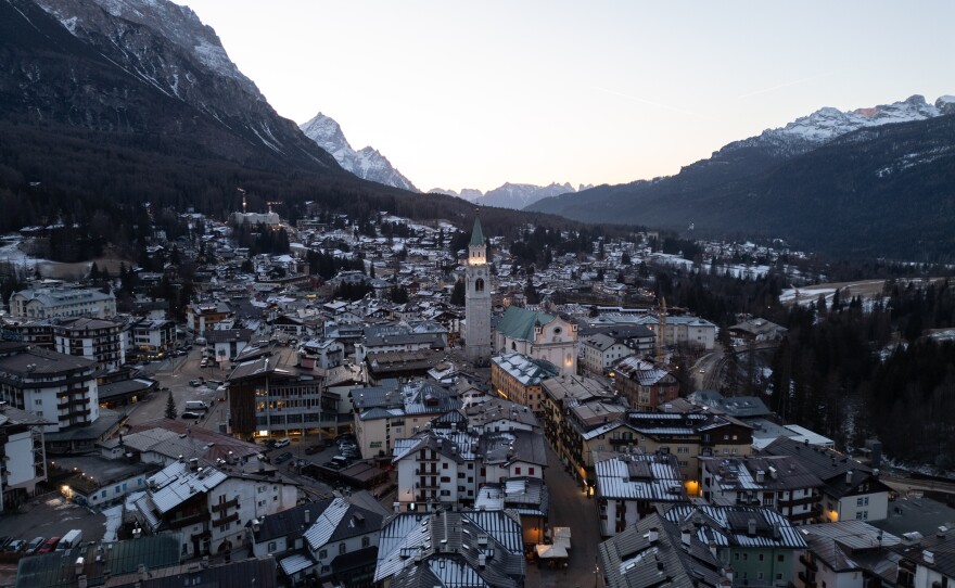The illuminated bell tower of the Basilica Minore dei Santi Filippo e Giacomo stands at the heart of Cortina d'Ampezzo, Italy, as evening settles over the valley. Once a small village of farmers and shepherds, this storied town has evolved into the "Pearl of the Dolomites," a renowned luxury destination. Surrounded by the limestone peaks of the UNESCO World Heritage Dolomites, the town's historic center remains a "living room" for celebrities and high society.