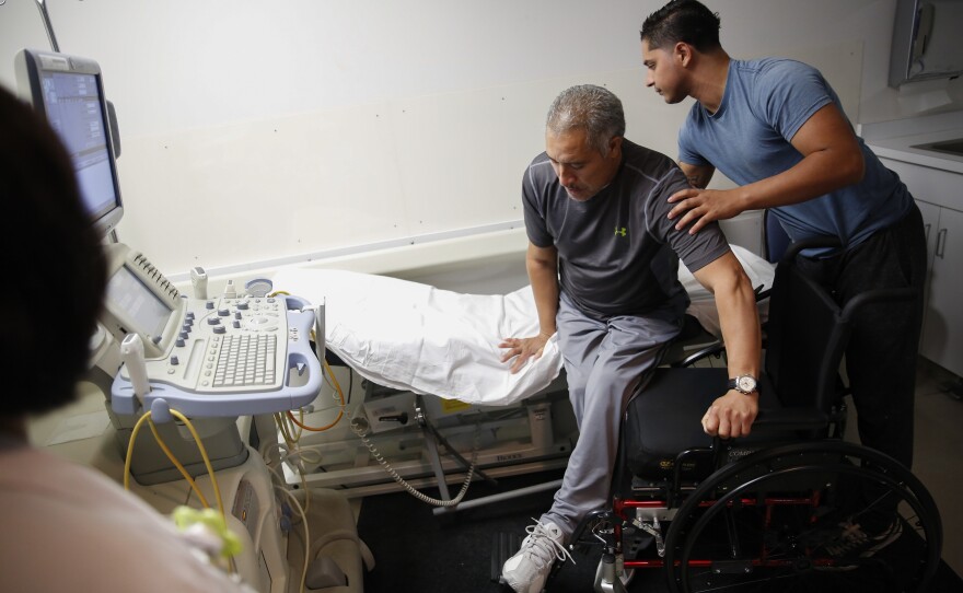 Joel Ramirez is helped onto a bed to receive an ultrasound with the aid of Francisco Guardado, his home health care giver, during a visit to the hospital in Rialto, Calif.