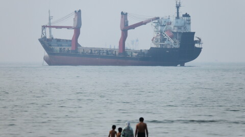 A group of people stands in shallow water as a cargo ship appears anchored in the Strait of Hormuz off Bandar Abbas, Iran, Tuesday, June 30, 2026.