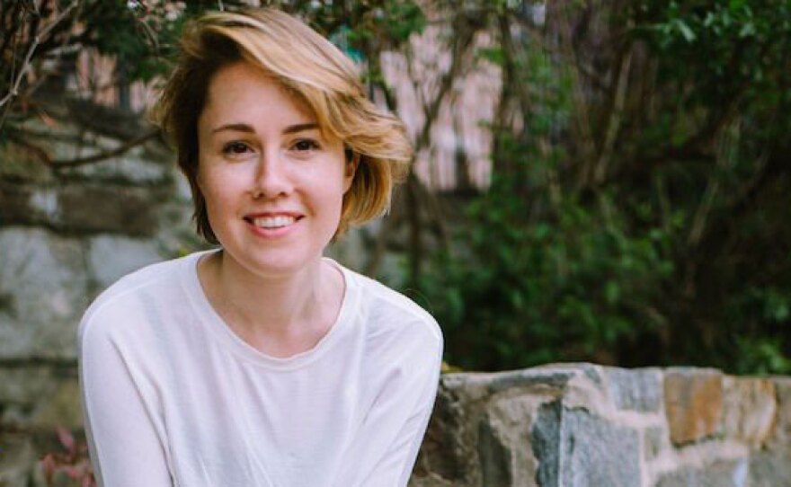 Composer Caroline Shaw sits near a garden wall, smiling and looking at the camera. She has short, light brown hair and light skin, and wears a white t-shirt.