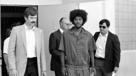 Kevin Cooper, center, a suspect in connection with the slashing death of four people in Chino, is escorted to a car for transport to San Bernadino from Santa Barbara, Calif., after he was arrested by police at Santa Cruz Island, July 31, 1983. 