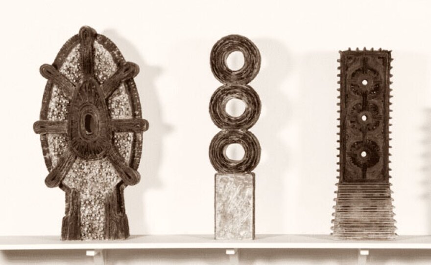 Three sculptures are shown on a shelf. Each is a gray-ish, taupe color that evokes steel.