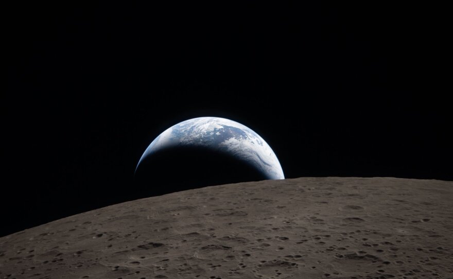 Earthset captured through the Orion spacecraft window at 6:41 p.m. ET on April 6 during the Artemis II crew's flyby of the moon. A muted blue Earth with bright white clouds sets behind the cratered lunar surface. The dark portion of Earth is experiencing nighttime. In the foreground, Ohm crater has terraced edges and a flat floor interrupted by central peaks. Central peaks form in complex craters when the lunar surface, liquefied on impact, splashes upward during the crater's formation.