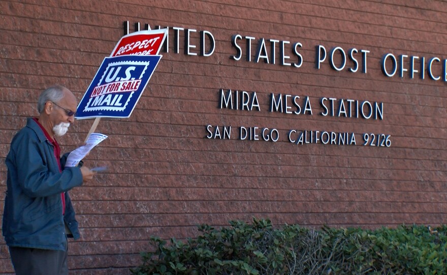 A postal worker is shown holding a sign that says "US mail not for sale" outside the Mira Mesa post office on March 20, 2025.