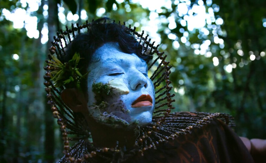 Occupying the center of the frame, a close up of a person whose face has been painted with blue make up and red lipstick and wears a headdress made of wood and twine. Their eyes and mouth are closed and they are surrounded by the lush green forest with dappled light streaming in.