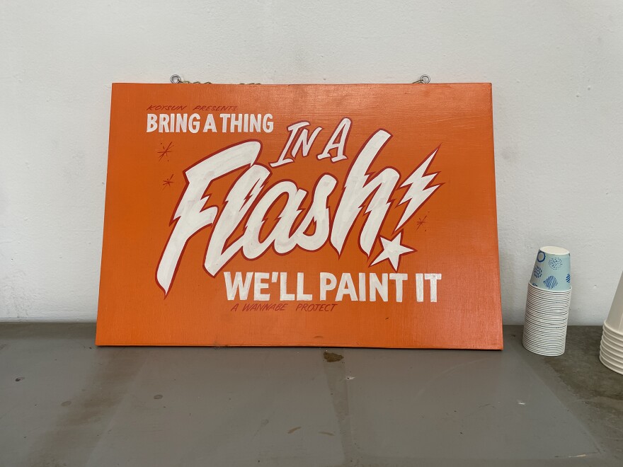 A hand-painted sign by San Diego artist Koy Sun emphasizes the generosity inherent in his sign painting. It is an orange wooden sign, propped against the wall. It reads "Bring a thing. In a FLASH! We'll paint it" in white lettering.