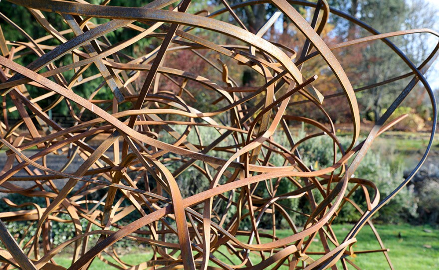"Bramble" (2024) by Matt Devine is shown in an undated photo. It's a scrambled, tangled large steel sculpture with a reddish-orange patina, situated outside in a grassy, wooded setting.